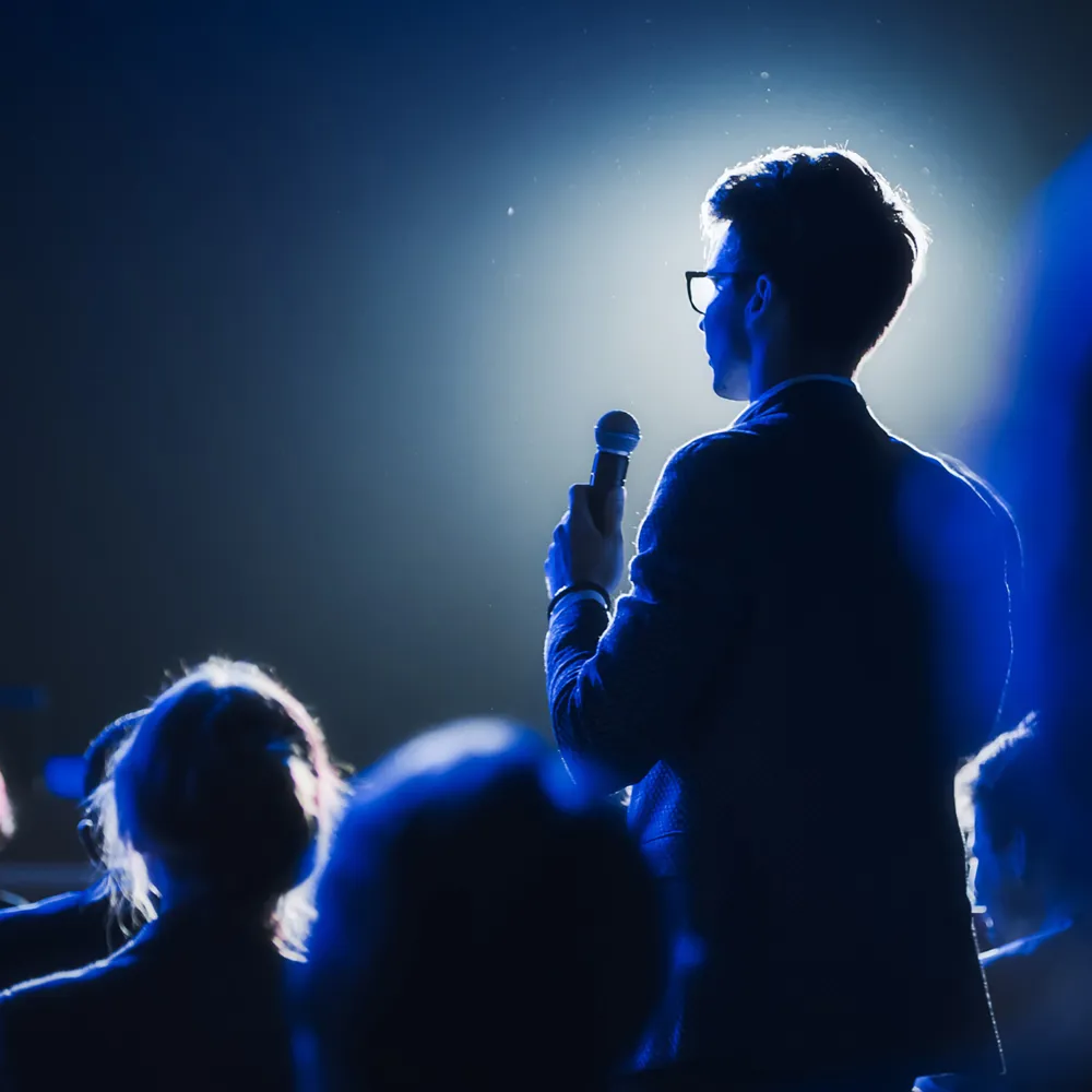 ALT Text: a man standing up in a crowd getting ready to ask a question during a keynote 